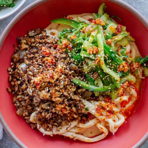 Bowl of chewy udon noodles coated in creamy peanut sauce, topped with crispy shredded tofu, cucumber-cilantro tiger salad, sesame seeds, and a drizzle of chili oil.