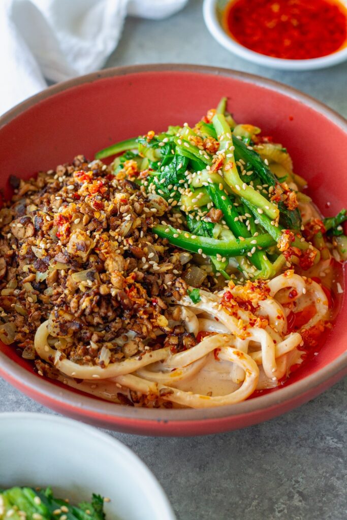 Bowl of chewy udon noodles coated in creamy peanut sauce, topped with crispy shredded tofu, cucumber-cilantro tiger salad, sesame seeds, and a drizzle of chili oil.
