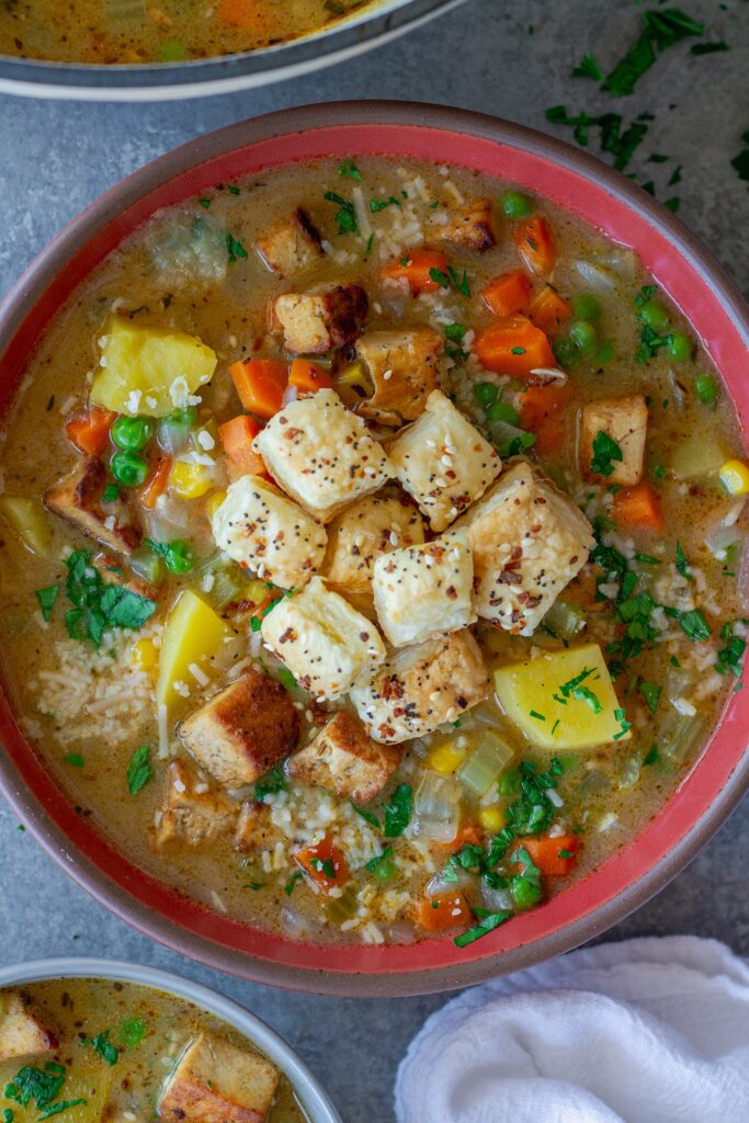 Bowl of creamy vegan chicken pot pie soup topped with crispy tofu, peas, and golden puff pastry croutons, garnished with parsley and parmesan