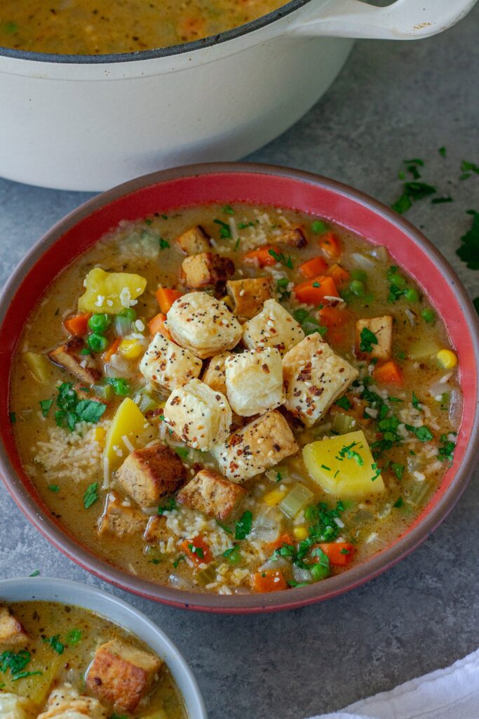 Bowl of creamy vegan chicken pot pie soup topped with crispy tofu, peas, and golden puff pastry croutons, garnished with parsley and parmesan