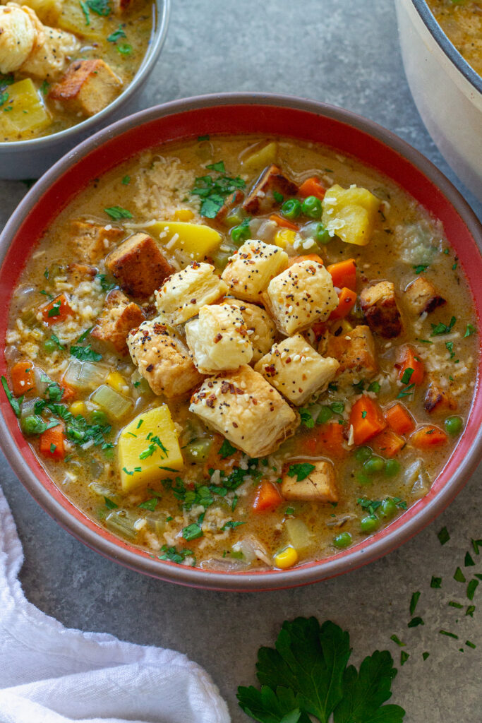 Bowl of creamy vegan chicken pot pie soup topped with crispy tofu, peas, and golden puff pastry croutons, garnished with parsley and parmesan